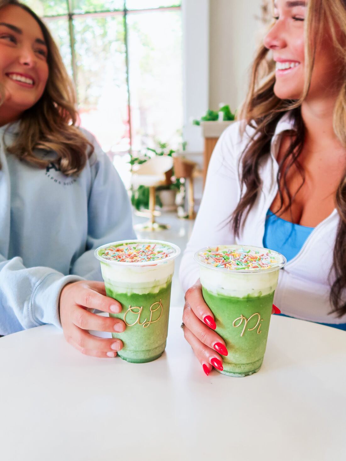 Two Alpha Delta Pi members laughing over matcha drinks with gold AD and Pi lettering