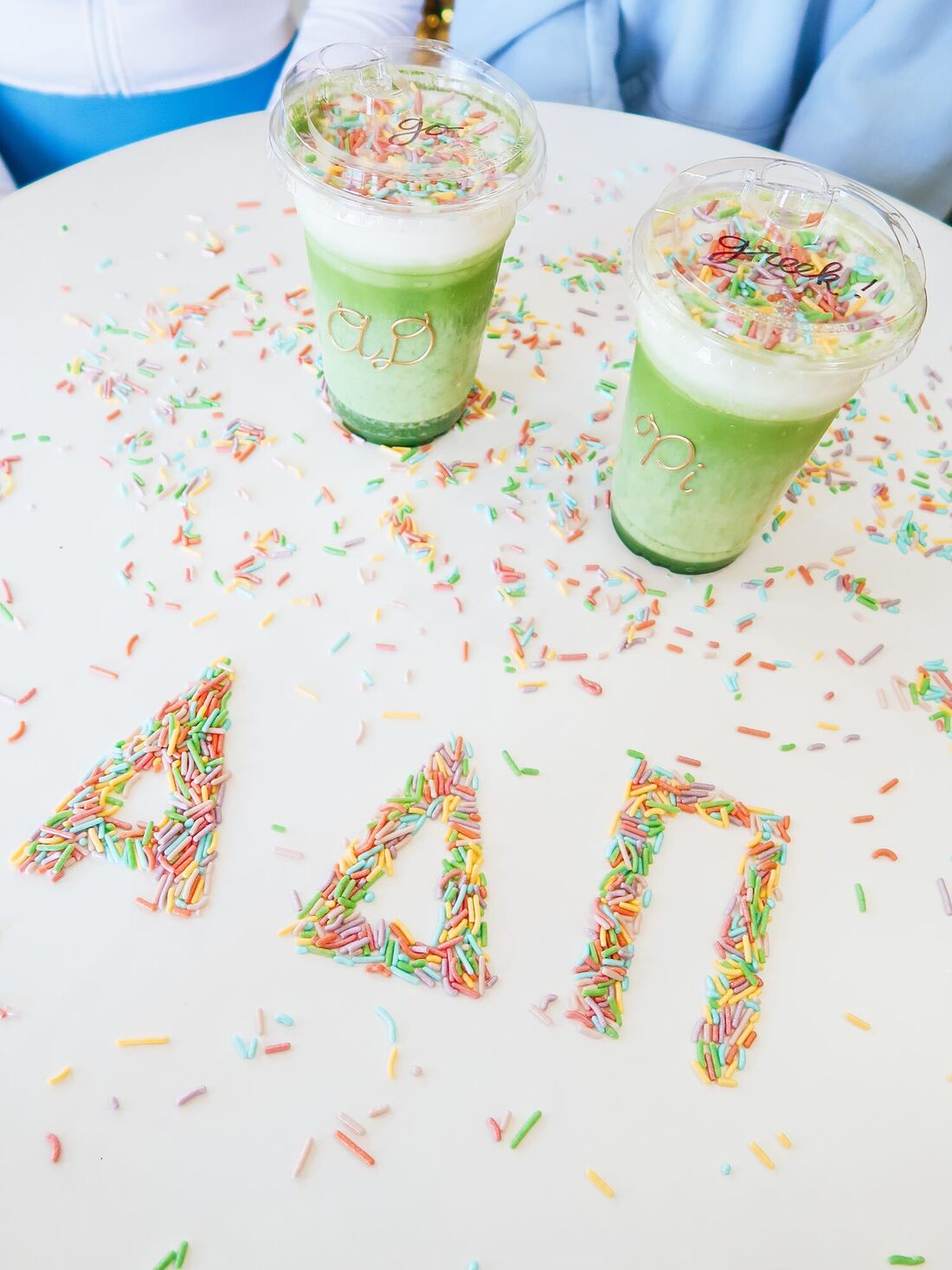 Two matcha drinks with gold AD and Pi lettering beside rainbow sprinkles spelling ΑΔΠ